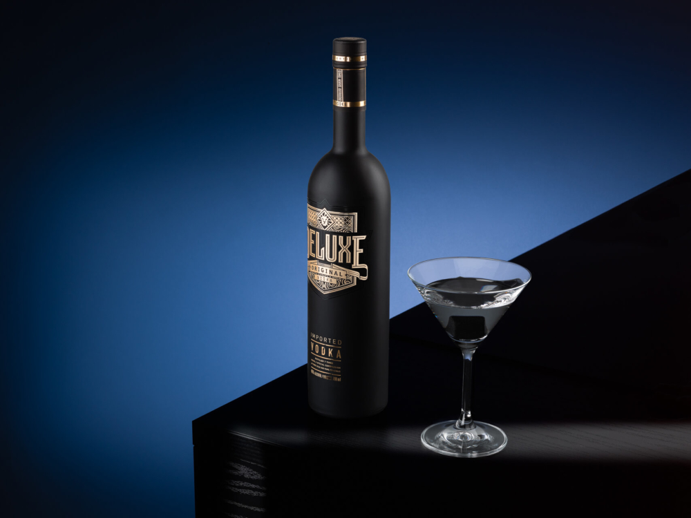 Une bouteille de vodka noire brillante d'une marque haut de gamme se trouve à côté d'un verre à martini rempli d'une boisson claire. Ils sont sur une surface noire brillante avec un fond bleu foncé.