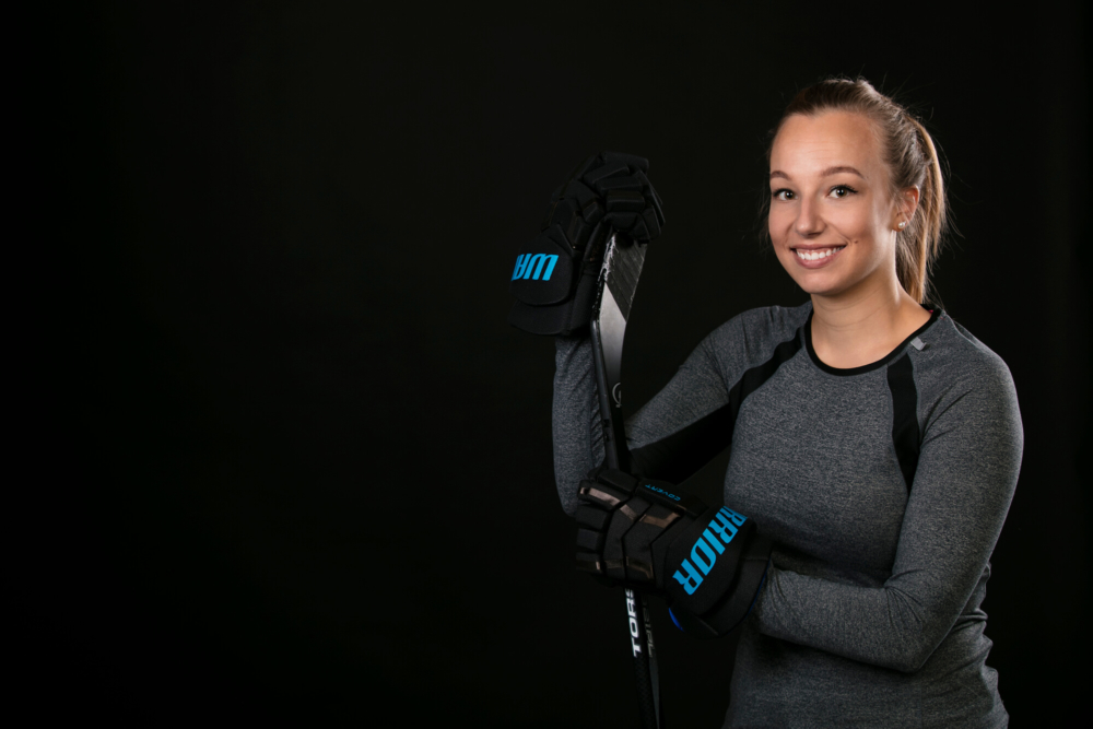 Une jeune femme heureuse portant des vêtements de sport porte un bâton de hockey sur glace sur son épaule sur un fond sombre.