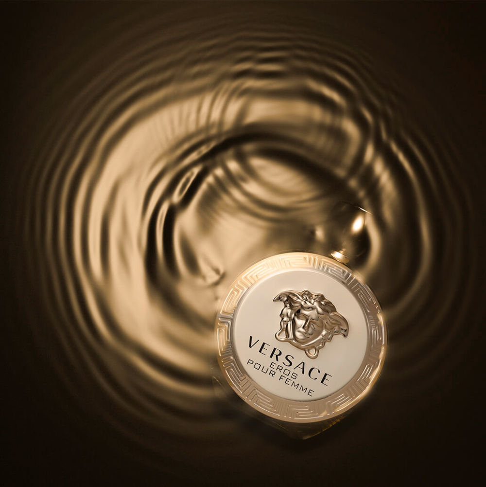 Vue d'en haut d'un bouchon doré de bouteille de parfum Versace, reflété dans de l'eau ondulante sur fond sombre.