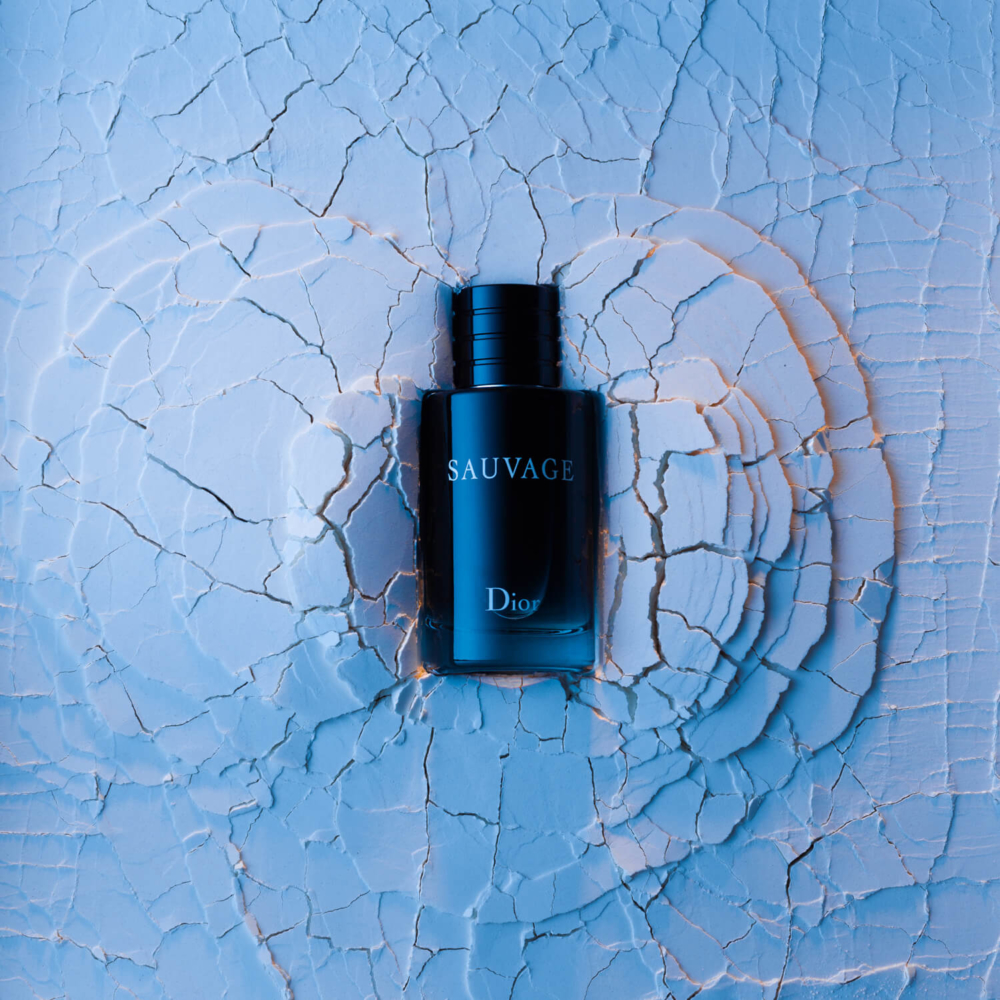 Une bouteille d'eau de Cologne Dior Sauvage est posée sur une surface bleue à la texture de peinture craquelée, doucement éclairée.