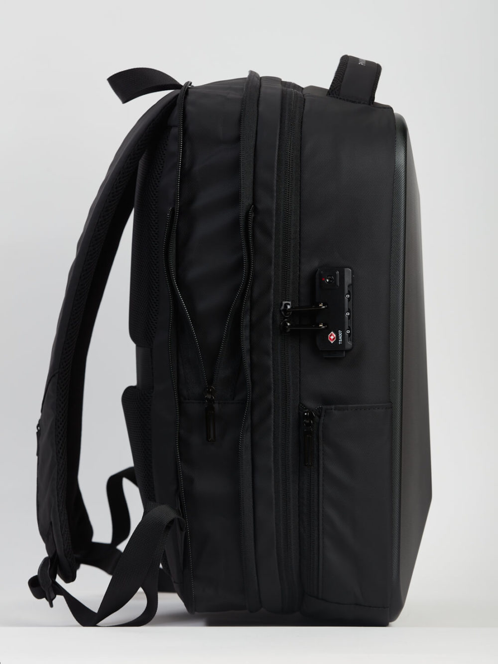 L'image montre un sac à dos noir avec de nombreuses poches et des bretelles rembourrées. Il possède une petite serrure à combinaison sur une poche latérale et est affiché sur fond gris.
