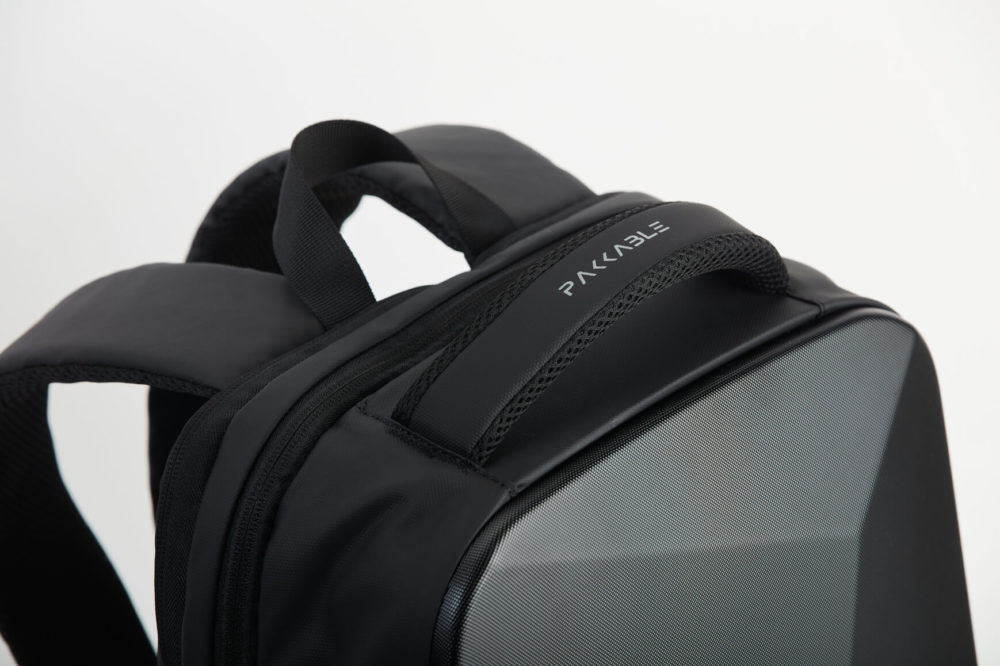 Un plan rapproché d'un sac à dos « raviel » noir et tendance est présenté. L'accent est mis sur le nom de la marque imprimé sur la poignée. L'extérieur texturé du sac à dos se détache sur un fond blanc.