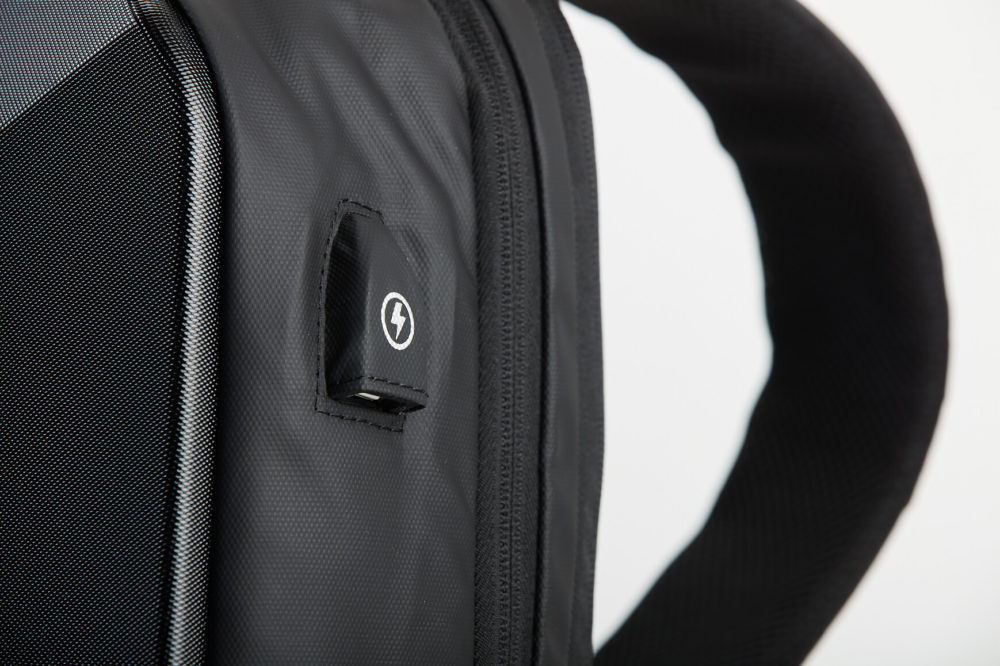 Un plan rapproché d’un sac à dos noir est montré. Il présente une surface texturée, un logo sur une petite étiquette en tissu et une partie d'une bandoulière rembourrée. Le fond est blanc.