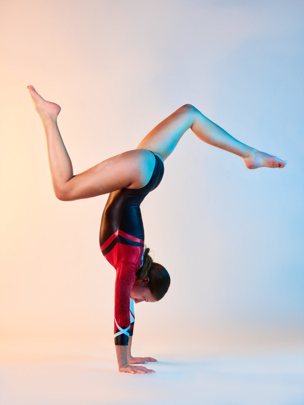 photographe sport yoga bien-être fitness danse slika photography 001 Une femme portant un justaucorps rouge et noir fait un poirier complexe sur un fond couleur pêche. Une de ses jambes est pliée et l’autre pointe vers le haut.
