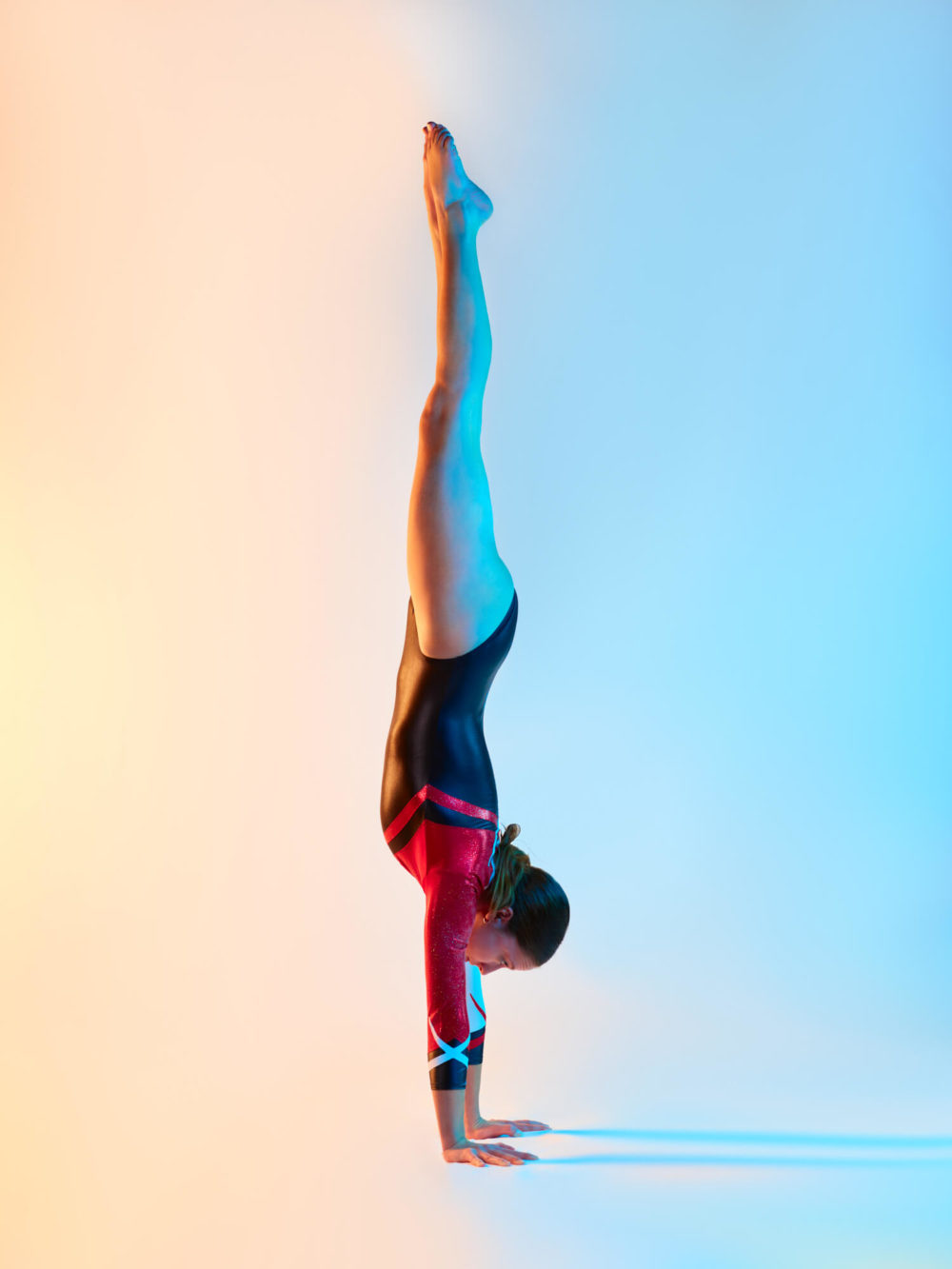 photographe sport yoga bien-être fitness danse slika photography 003 Une femme, vêtue de vêtements de sport noirs et rouges, fait le poirier sur un fond qui passe du bleu à l'orange. Son corps est droit, montrant son équilibre et sa puissance.