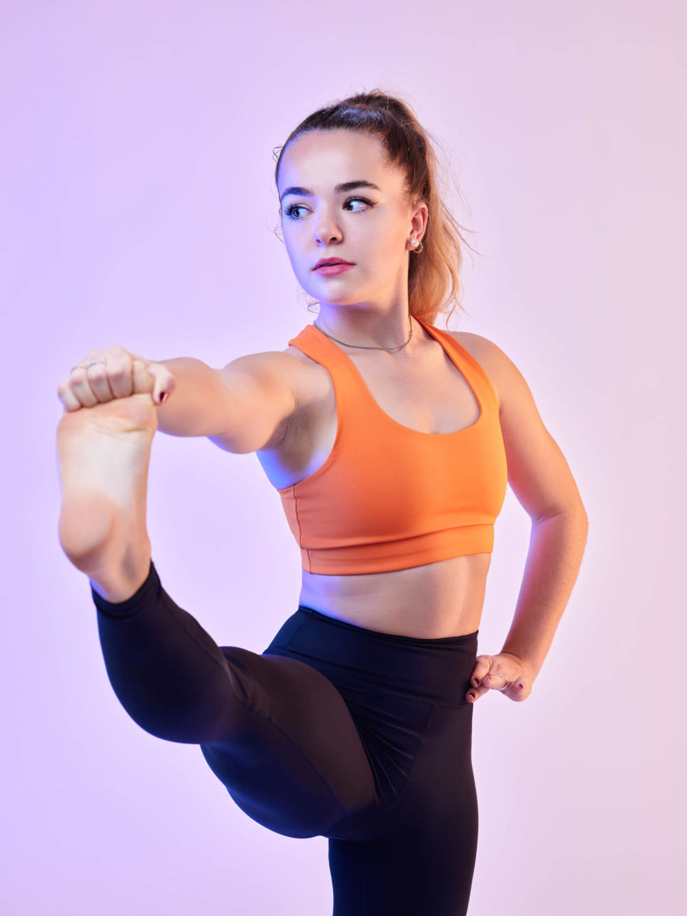 photographe sport yoga bien-être fitness danse slika photography 007 Une femme concentrée et déterminée portant un soutien-gorge de sport orange et des leggings noirs fait un high kick. Elle est placée sur un fond dégradé rose et violet.