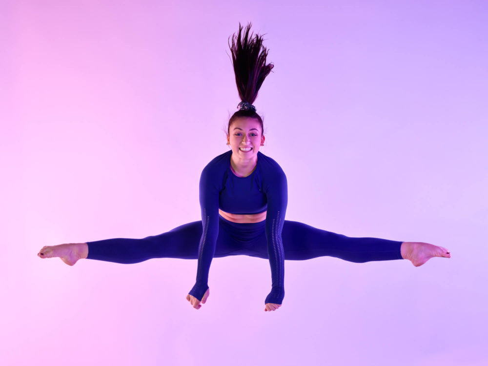 photographe sport yoga bien-être fitness danse slika photography 008 Une gymnaste vêtue d'une tenue bleue et noire fait un saut divisé en l'air. Elle est sur un fond rose et violet. Ses cheveux sont attachés haut et elle a l'air heureuse.