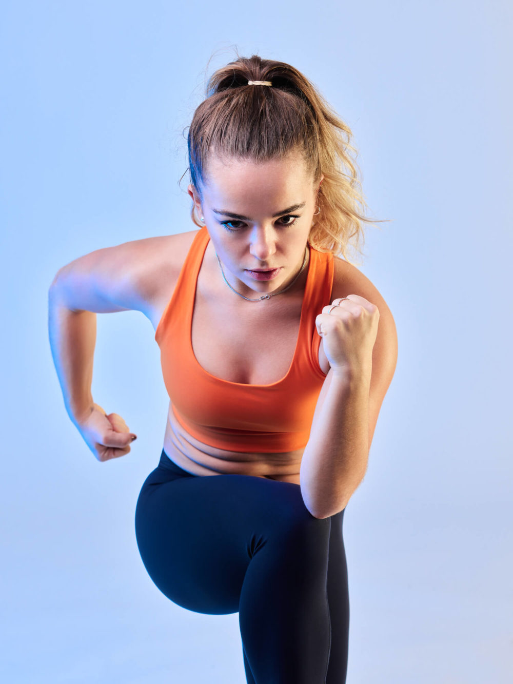 photographe sport yoga bien-être fitness danse slika photography 011 Une femme portant un soutien-gorge de sport orange et des leggings noirs court sur un fond bleu clair. Elle a l'air concentrée et déterminée.