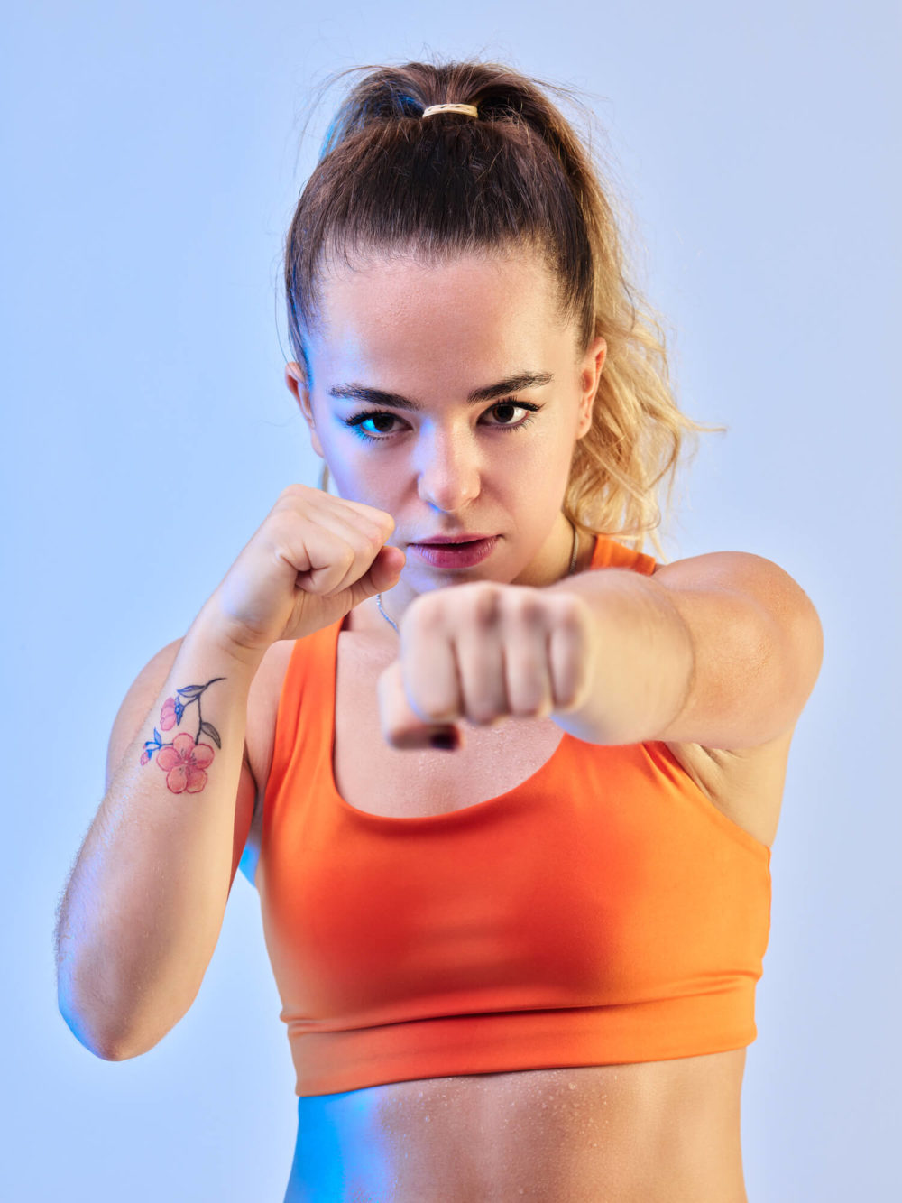 photographe sport yoga bien-être fitness danse slika photography 012 Une femme avec une queue de cheval et un tatouage sur le bras, vêtue d'un haut de sport orange, se dirige vers la caméra. Le fond est bleu clair.