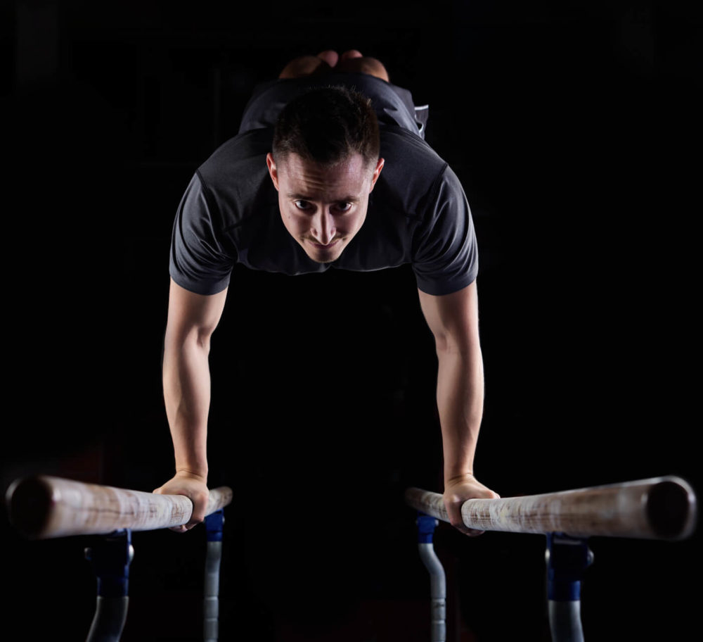 photographe sport yoga bien-être fitness danse slika photography 013 Un homme fait une routine de gymnastique aux barres parallèles, se concentrant et faisant des efforts, avec un fond sombre derrière lui.