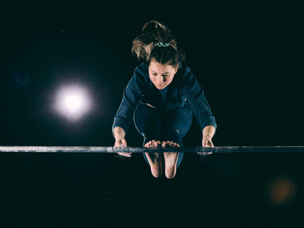 photographe sport yoga bien-être fitness danse slika photography 015 Une jeune gymnaste exécute une routine à la poutre. Elle est au milieu d'un saut et mise en évidence par un projecteur sur un fond sombre, montrant sa concentration et ses capacités athlétiques.