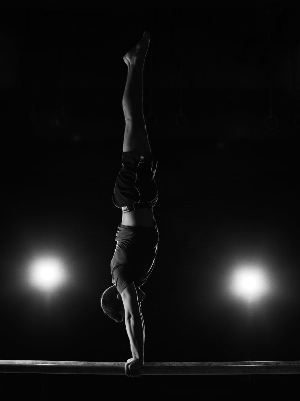 photographe sport yoga bien-être fitness danse slika photography 016 Un gymnaste fait le poirier sur une poutre, éclairé par des lumières frappantes dans un décor sombre, démontrant sa puissance et son équilibre en noir et blanc.