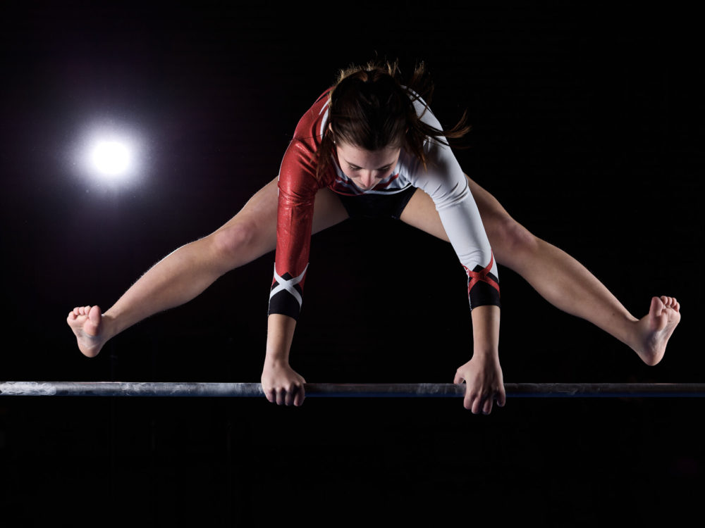 photographe sport yoga bien-être fitness danse slika photography 017 Une gymnaste fait un grand saut par-dessus une poutre dans une salle de sport faiblement éclairée, avec une lumière vive brillant derrière elle. Elle porte un justaucorps rouge et noir.