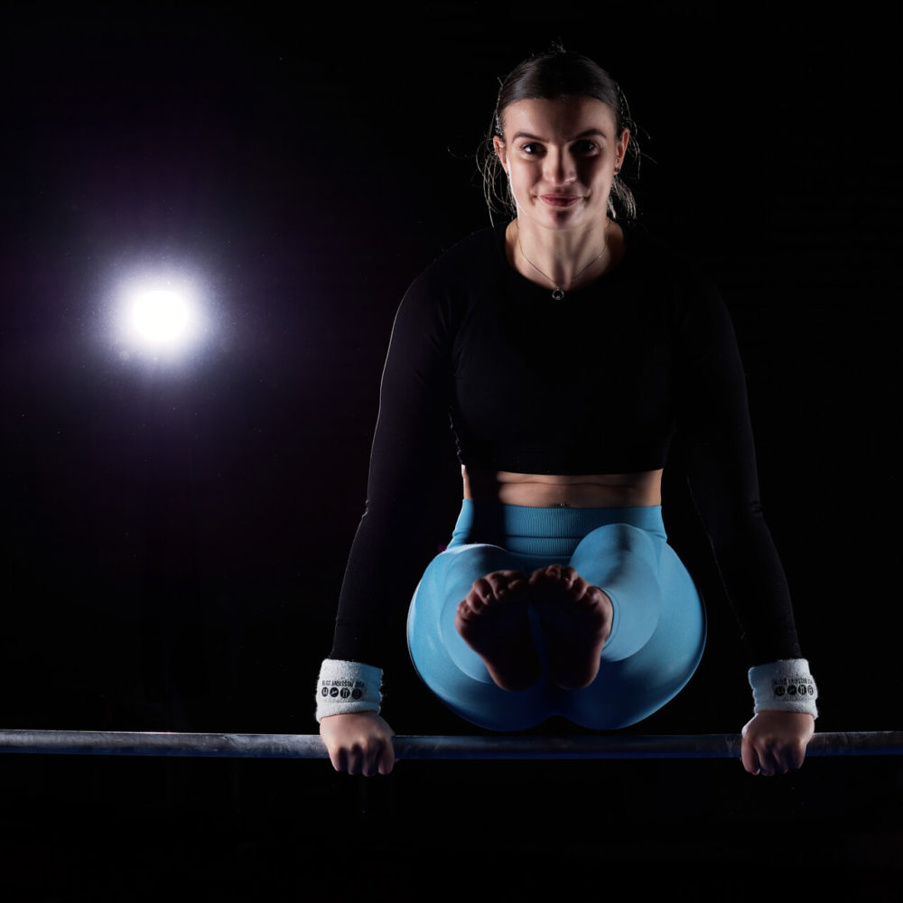 photographe sport yoga bien-être fitness danse slika photography 018 Une gymnaste vêtue d'un haut noir et de leggings bleus tient la poutre avec un projecteur qui brille par derrière, ce qui souligne son aspect concentré et sa position confiante.