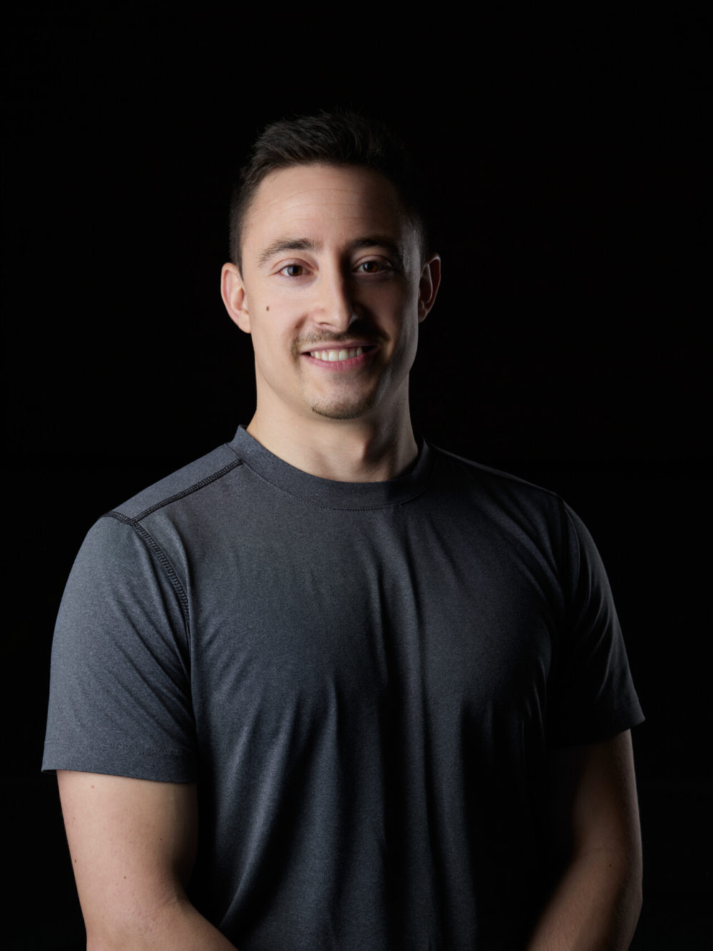 photographe sport yoga bien-être fitness danse slika photography 019 Une photo montre un homme aux cheveux courts souriant. Il porte un t-shirt gris foncé et se tient sur un fond noir, regardant droit dans la caméra.