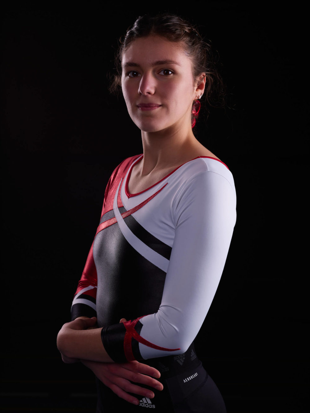 photographe sport yoga bien-être fitness danse slika photography 022 Une gymnaste vêtue d'un justaucorps blanc et rouge se tient en toute confiance, les bras croisés sur un fond sombre. Elle porte des boucles d'oreilles rouges et a l'air déterminée.