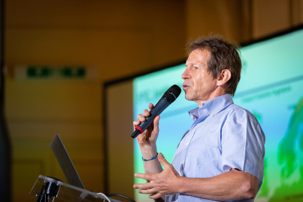 Un homme prend la parole lors d’une conférence, utilise un microphone et se tient à côté d’un ordinateur portable. Une diapositive de présentation est visible en arrière-plan.