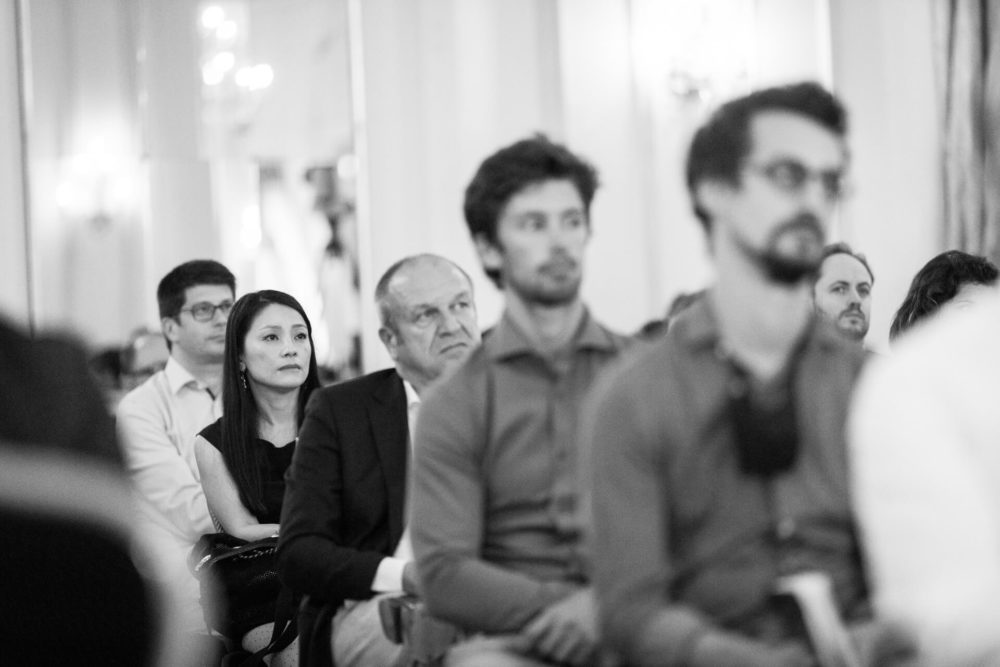 Une simple photo en noir et blanc montre un groupe diversifié d’adultes attentifs assis en rangées lors d’une conférence ou d’une prise de parole en public. L'arrière-plan est un peu flou tandis que le premier plan est plus net.