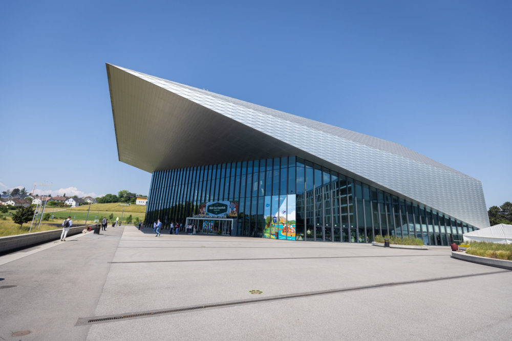 Un bâtiment moderne avec une façade en verre et un toit métallique unique accueille les visiteurs sous un ciel bleu clair. Une grande zone pavée mène à l'entrée, entourée d'un paysage herbeux.