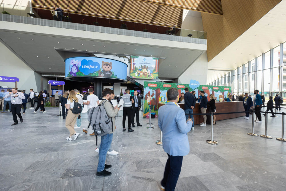 Des gens assistent à une conférence dans un grand hall doté de stands promotionnels Salesforce. La zone est bondée de participants qui réseautent et regardent autour d'eux.