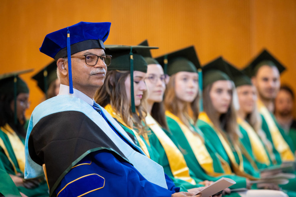 Un professeur d'âge moyen, portant des lunettes et une tenue de remise des diplômes, est assis devant un groupe de jeunes diplômés en robes vertes et jaunes lors d'une cérémonie de remise des diplômes.