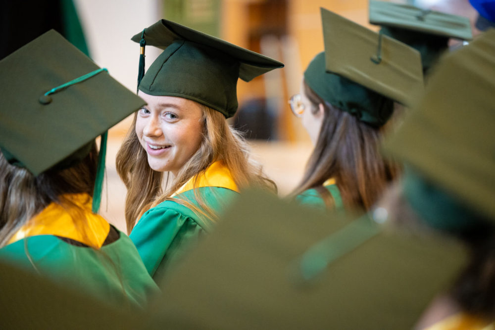 Une jeune femme souriante vêtue d'une tenue de remise des diplômes verte et dorée se retourne parmi un groupe d'autres diplômés.