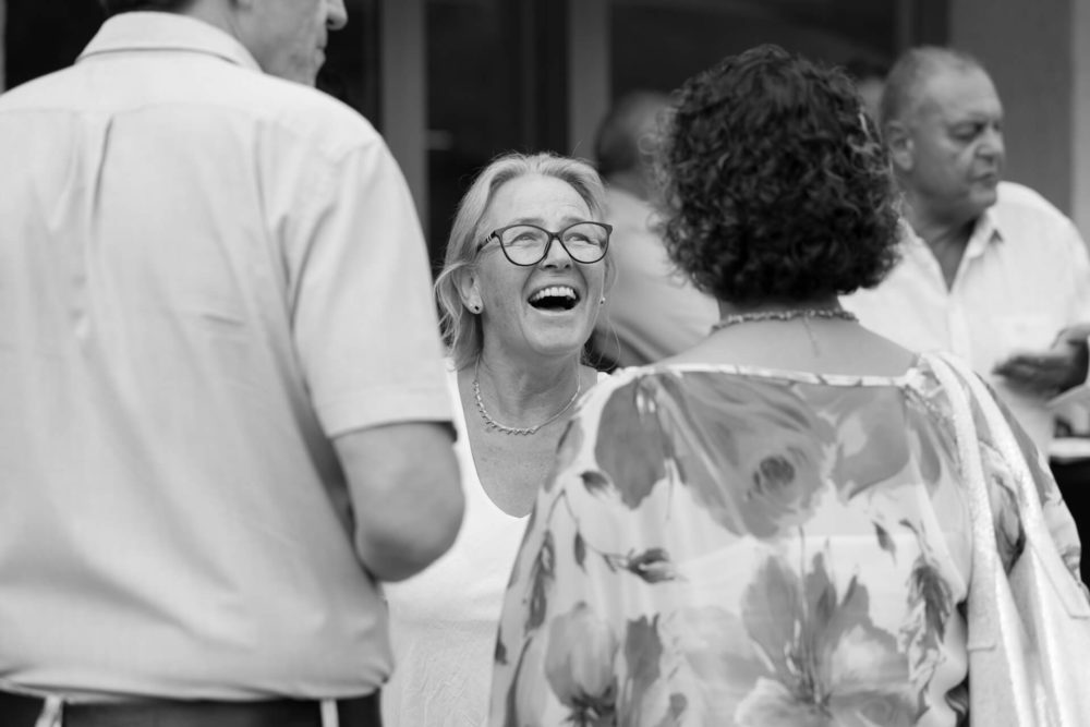 Une vieille femme heureuse portant des lunettes, riant bruyamment tout en parlant à des amis lors d'un événement social, représentée en noir et blanc.