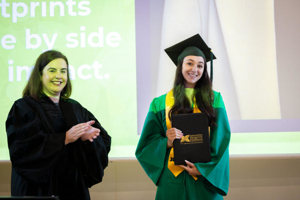Une femme récemment diplômée porte des vêtements verts et possède un diplôme. Elle se tient à côté d’une autre femme souriante et vêtue d’une tenue noire de remise des diplômes. Ils sont devant un arrière-plan qui dit « les empreintes côte à côte ont un impact.