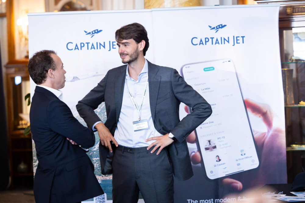 Deux hommes discutent lors d'un événement professionnel près d'un stand « Captain Jet » avec des banderoles promotionnelles. Un homme fait des gestes tandis que tous deux sourient.