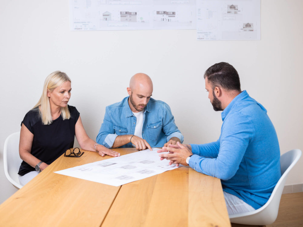 Trois experts, une femme et deux hommes, discutent de conceptions architecturales autour d'une table dans un bureau bien éclairé aux murs blancs.
