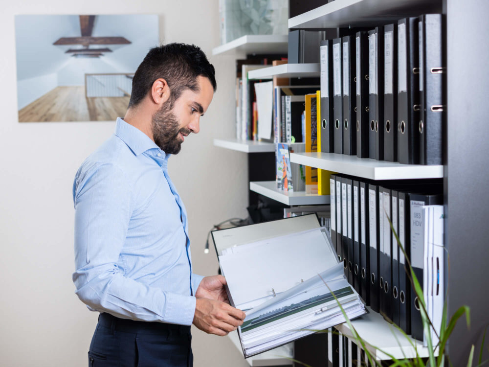 Un homme en chemise bleue vérifie des fichiers dans un dossier tout en se tenant à côté d'une étagère remplie de dossiers dans un bureau contemporain.