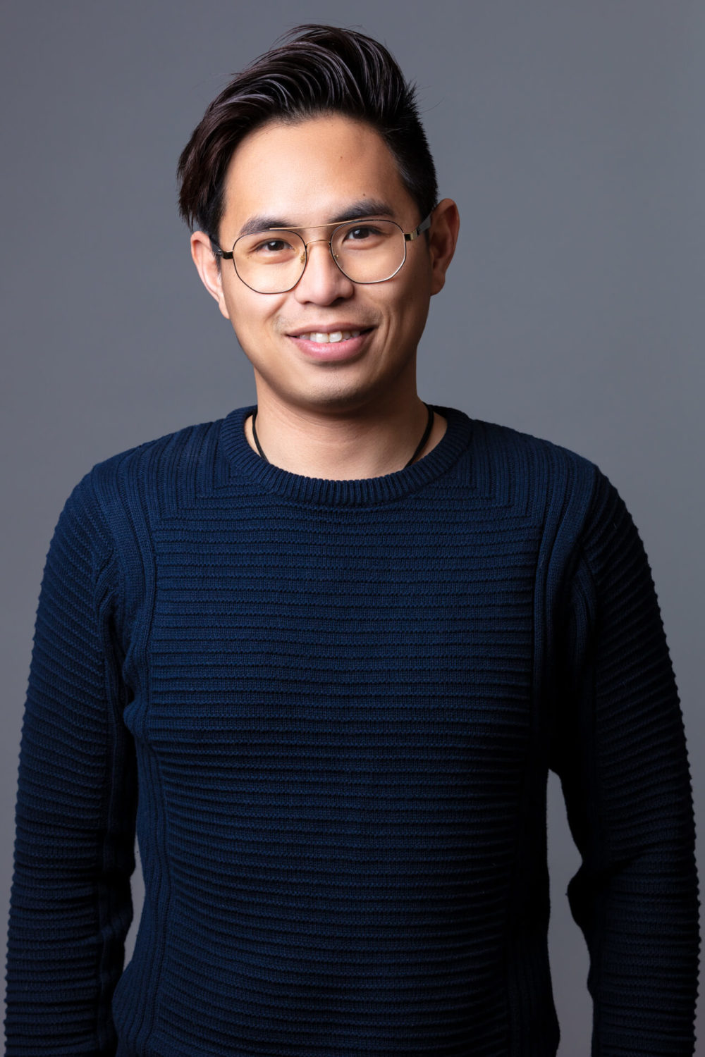 Un jeune homme asiatique heureux avec des lunettes et un pull bleu foncé se tient devant un fond gris.