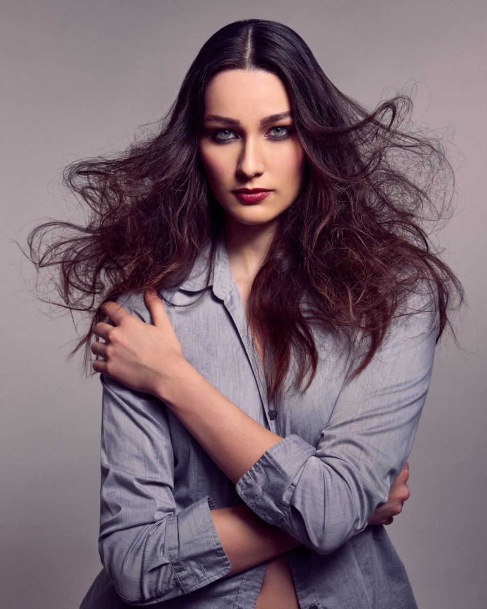 Shooting_photo_Portrait_Studio_Séance_Emotion_Artistique_027 Une femme aux cheveux bruns et aux yeux bleus porte un chemisier gris. Elle a les bras croisés et regarde directement la caméra.