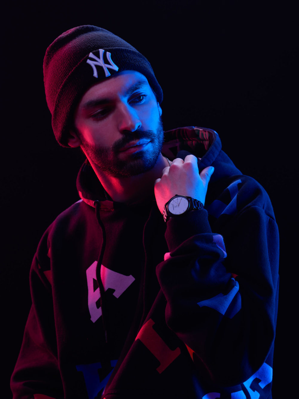Shooting_photo_Portrait_Studio_Séance_Emotion_Artistique_036 Un gars portant un bonnet et un sweat à capuche, éclairé par des lumières bleues et rouges, positionne pensivement sa main près de son menton pour montrer sa montre.