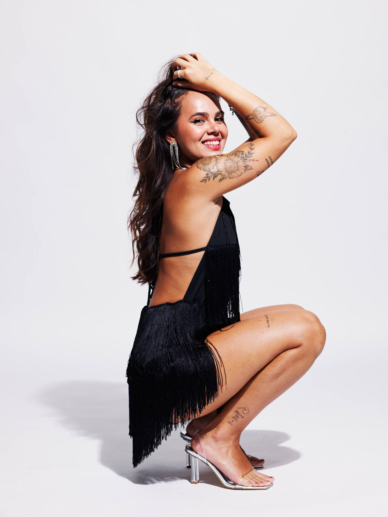Shooting_photo_Portrait_Studio_Séance_Emotion_Artistique_051 Une femme tatouée et aux longs cheveux noirs sourit et pose dans une robe à franges noire avec des talons argentés. Elle touche ses cheveux de manière ludique sur un fond blanc.