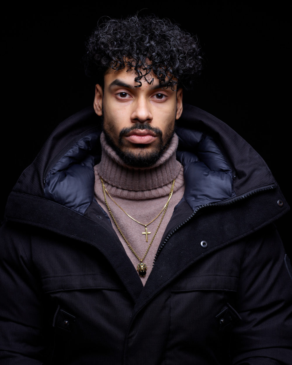 CF018735 Portrait d'un homme aux cheveux bouclés, vêtu d'une veste noire et d'un pull beige. Il arbore une expression concentrée et porte un collier avec une croix en or. Un fond noir le met en valeur.