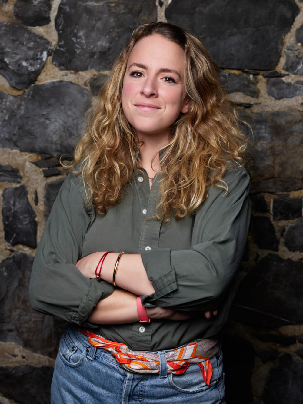 Une femme aux cheveux longs et bouclés sourit doucement face à l'appareil photo, les bras croisés. Elle porte une chemise verte et un jean bleu, se tenant devant un fond de mur en pierre sombre.