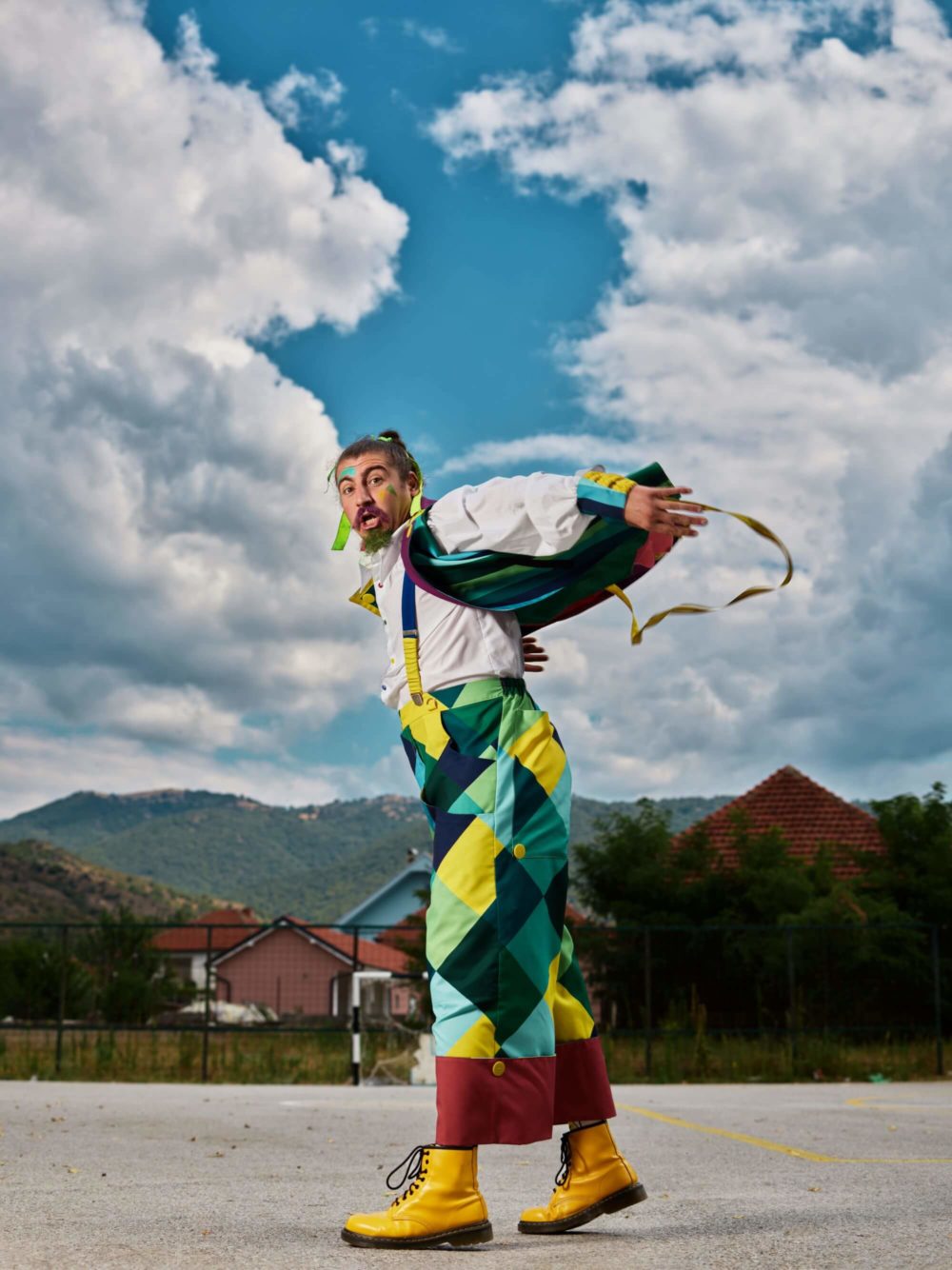 Kole Kitanov by Kre Ivanov Un homme portant une salopette colorée et une chemise blanche prend une pose amusante en plein air. Il a une moustache et un air surprise. Le ciel est partiellement nuageux, avec des collines et des maisons en arrière-plan.