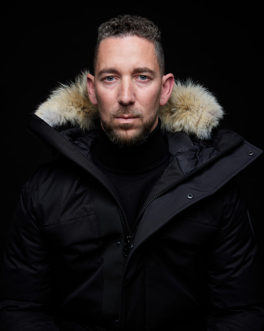 P0015570 Un homme à la barbe courte, regardant droit dans l'appareil photo, porte un manteau noir avec une doublure en fourrure sur la capuche, sur fond sombre.