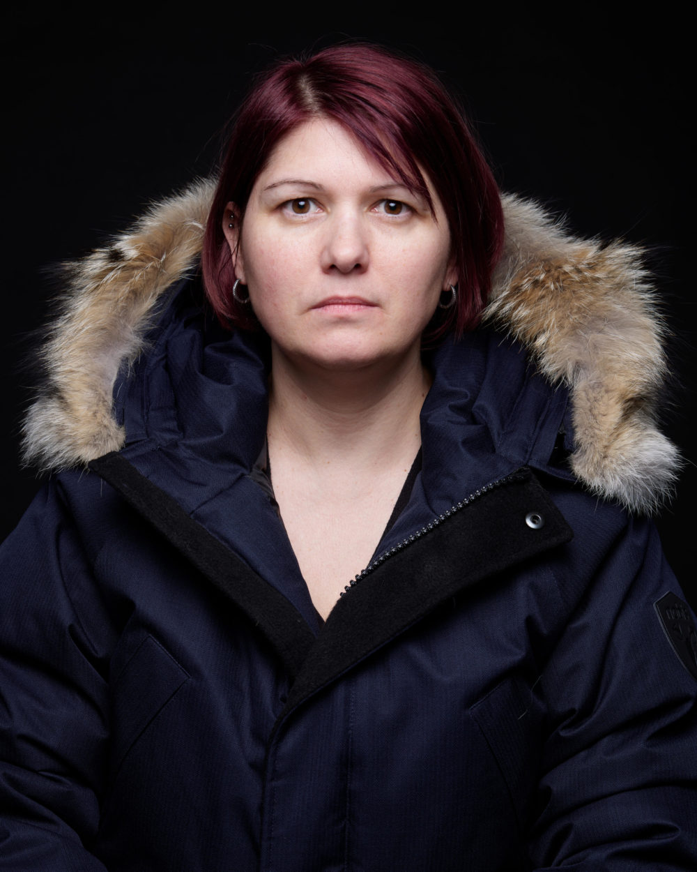 P0015733 Une femme aux cheveux courts teints en rouge et à l'expression sérieuse, portant une veste d'hiver noire avec capuche bordée de fourrure, sur un fond noir.