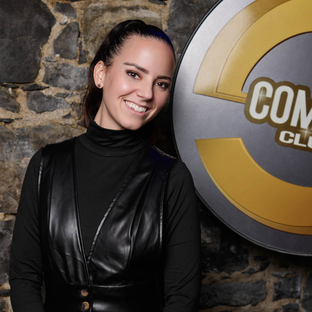 Priscilla Formaz au Swiss Comedy Club - Kire Ivanov Une femme souriante avec une queue de cheval, portant un col roulé noir et un gilet en cuir, se tient devant un mur de pierre arborant le grand emblème