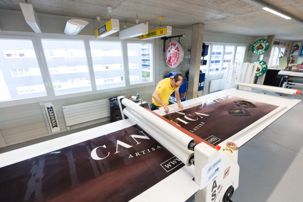 Deux personnes travaillent dans une imprimerie, manipulant une imprimante grand format pour imprimer une bannière en vinyle. Elles sont entourées de divers outils et fournitures d'impression.