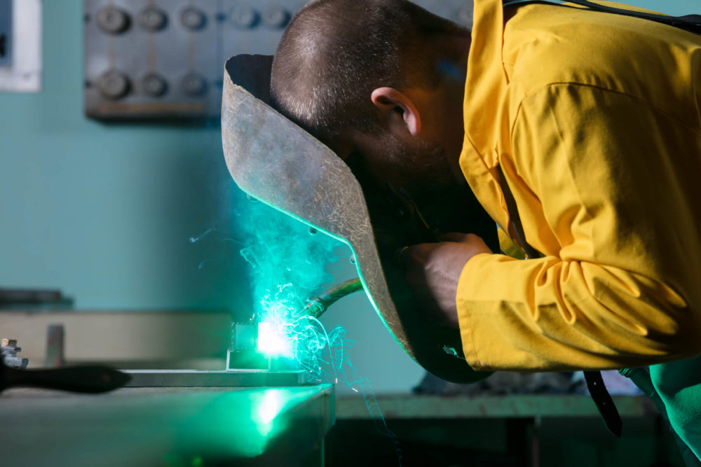 Un ouvrier qualifié en veste jaune soudure du métal en utilisant un équipement de protection, des étincelles vertes brillantes jaillissantes dans un atelier peu éclairé.